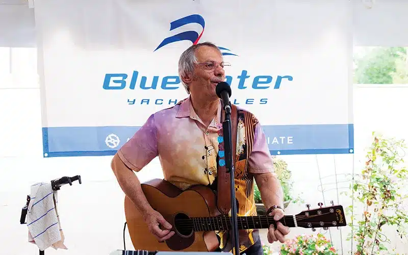 An elderly man in a colorful shirt plays an acoustic guitar and sings into a microphone.