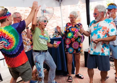 A group of older adults wearing colorful, tie-dye clothing and headbands are celebrating and laughing under a tent.